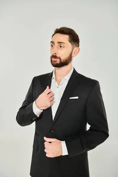 Hombre de negocios barbudo elegante pensativo en traje negro de pie y mirando hacia otro lado en el fondo gris - foto de stock
