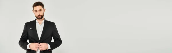 Charismatic bearded businessman buttoning black suit and looking at camera on grey, banner — Stock Photo