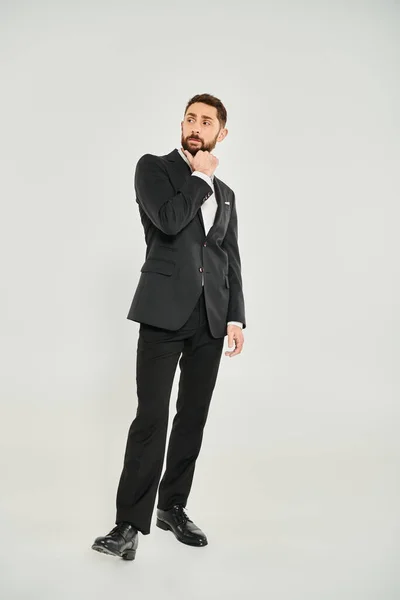 Full length of thoughtful businessman in black suit touching beard and looking away on grey — Stock Photo