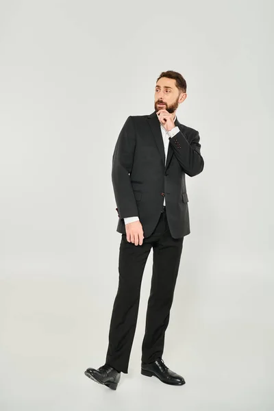 Full length of serious elegant businessman in black suit touching beard and looking away on grey — Stock Photo