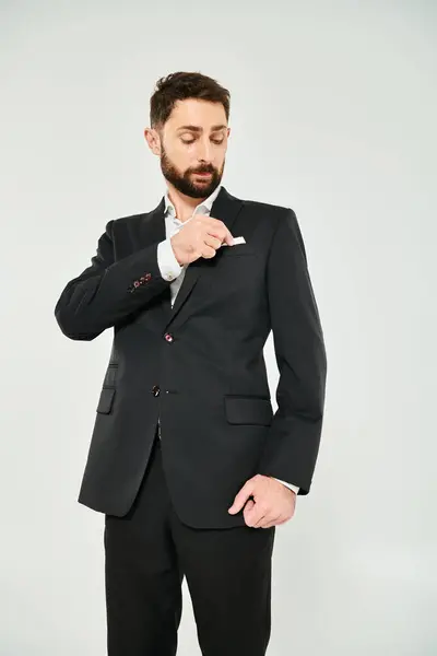 Elegante hombre de negocios barbudo ajustando pañuelo blanco en el bolsillo del pecho de chaqueta negra en gris - foto de stock
