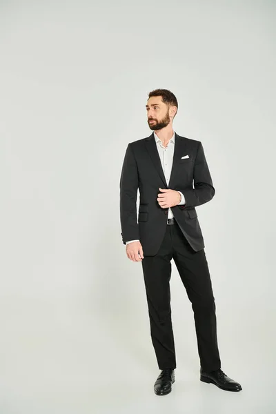 Повна довжина стильного бізнесмена в чорному елегантному костюмі, дивлячись далеко, стоячи на сірому — стокове фото