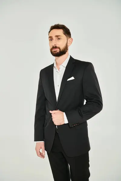 Carismático hombre de negocios barbudo en traje elegante negro mirando hacia otro lado mientras está de pie sobre fondo gris - foto de stock
