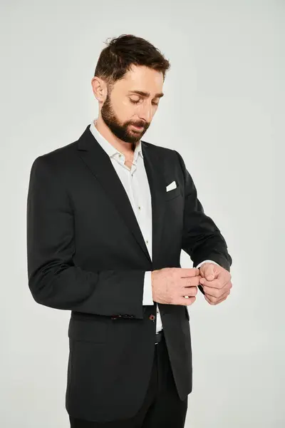 Hombre de negocios bien vestido ajustando la manga de traje elegante negro mientras está de pie sobre fondo gris - foto de stock