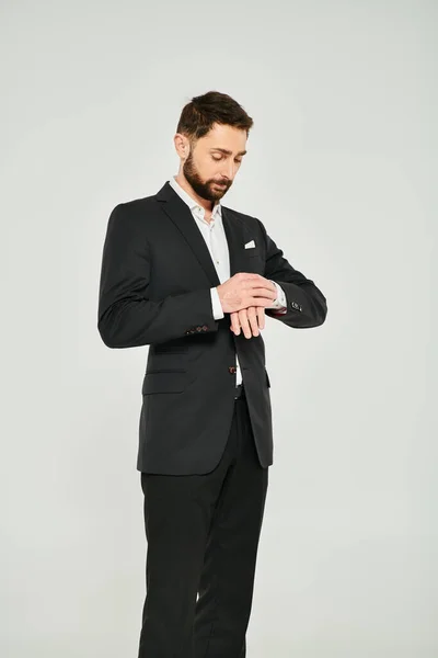 Гарний бородатий бізнесмен регулює рукав чорного костюма на сірому тлі в студії — стокове фото
