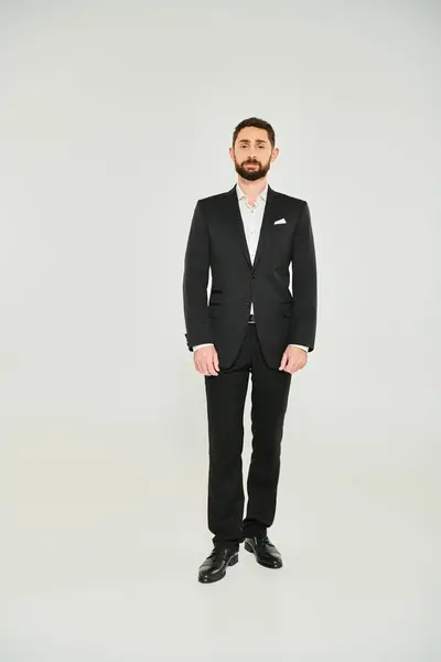 Front view of bearded businessman in black elegant suit standing on grey backdrop, full length — Stock Photo