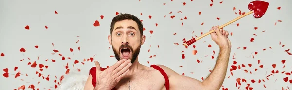 Homem cupido surpreso segurando flecha e cobrindo a boca aberta com a mão sob confete vermelho, bandeira — Fotografia de Stock