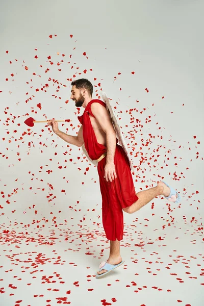 Hombre en traje de Cupido posando con flecha en forma de corazón bajo confeti rojo sobre gris, día de San Valentín - foto de stock