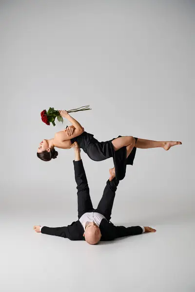 Mulher flexível em traje preto segurando rosas vermelhas e equilibrando em pés de parceiro de dança em cinza — Fotografia de Stock