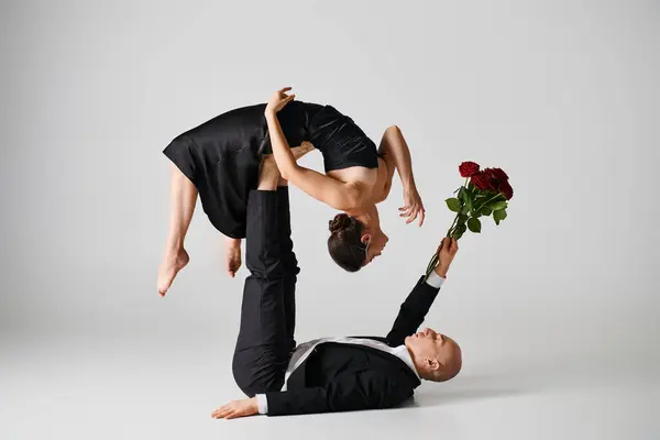 Mujer flexible en traje negro equilibrio en los pies de la pareja de baile sosteniendo rosas rojas en gris - foto de stock