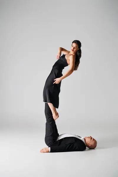 Гибкая молодая женщина в черном, балансирующая на босых ногах спортивного мужчины на сером фоне — стоковое фото