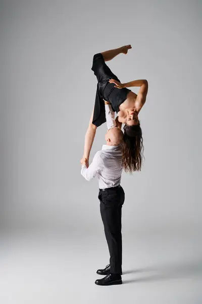 Starker Mann hebt brünette junge Frau in schwarzem Kleid, während er vor grauem Hintergrund tanzt — Stockfoto