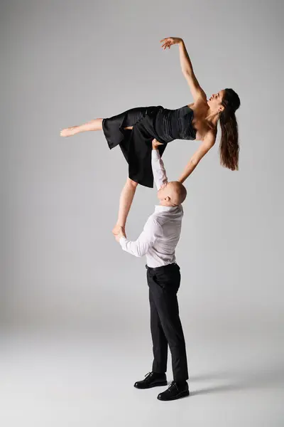 Homem forte levantando mulher morena flexível em vestido preto enquanto executa dança em pano de fundo cinza — Fotografia de Stock