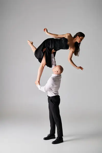 Сильний чоловік піднімає брюнетку молоду жінку в чорній сукні під час виконання танцю на сірому фоні — стокове фото