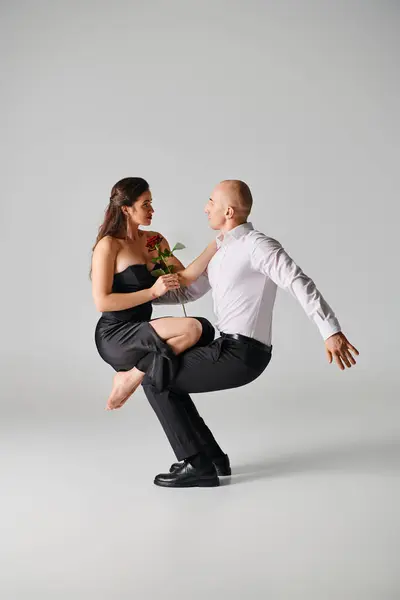 Brunette woman in black dress holding red rose and balancing on laps of male dancer on grey backdrop — Stock Photo