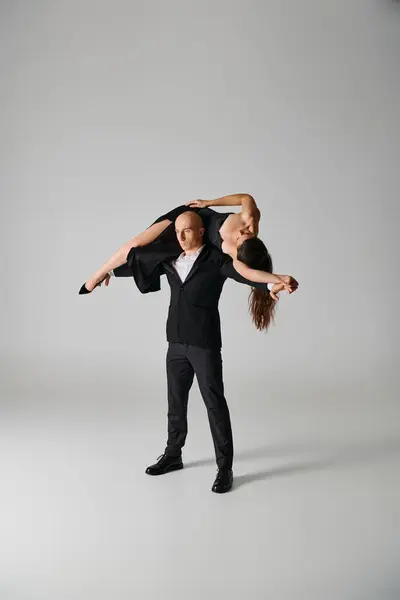 Сильный танцор, поднимающий брюнетку женщина в платье и на высоких каблуках на сером фоне в студии — стоковое фото