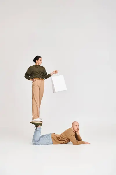 Mujer joven en ropa casual con bolsa de compras equilibrio en las piernas del hombre en una pose lúdica en el estudio - foto de stock