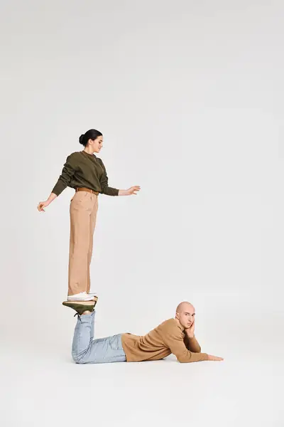 Young woman in casual wear balancing on legs of man in a playful pose in studio on grey backdrop — Stock Photo