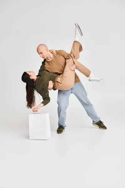 Man in casual attire holding in arms beautiful brunette woman with shopping bag on grey backdrop — Stock Photo