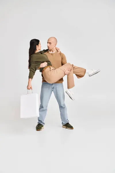 Hombre en traje casual sosteniendo en brazos hermosa mujer morena con bolsa de compras sobre fondo gris - foto de stock