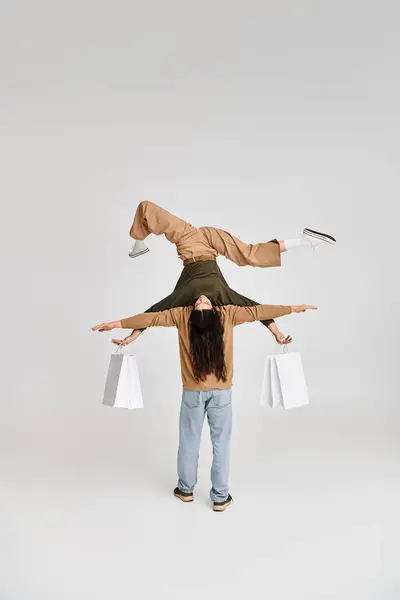 Акробатична жінка тримає покупки і балансує догори ногами за підтримки партнера на сірому — стокове фото