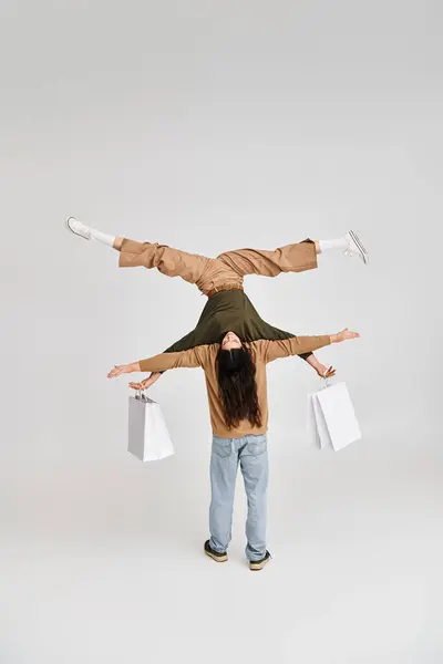 Жінка тримає сумки для покупок і балансує догори ногами за підтримки акробатичного партнера на сірому — стокове фото