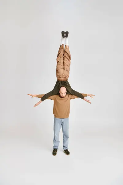 Strong man in casual wear supporting body of acrobat partner while rehearsing in grey studio — Stock Photo