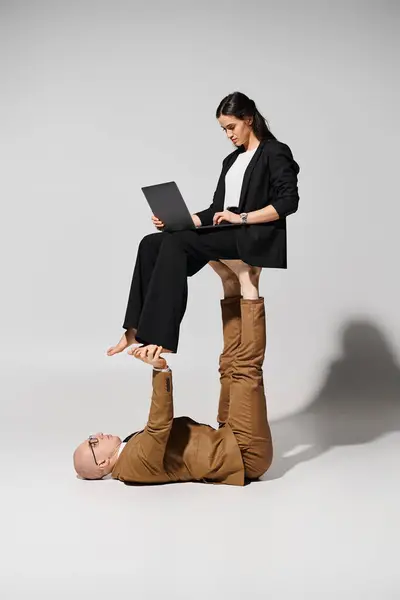 La mujer de negocios en el atuendo con el ordenador portátil que equilibra a los pies del hombre en el traje en gris, un par de acróbatas - foto de stock