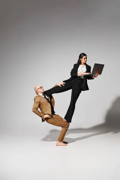 Frau in Businesskleidung mit Laptop balanciert mit Unterstützung des Mannes vor grauem Hintergrund, Akrobaten — Stockfoto