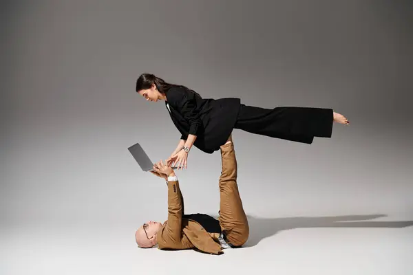 Woman in business wear with laptop balancing on feet of man in suit on grey, couple of acrobats — Stock Photo