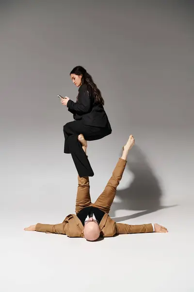 Femme d'affaires avec smartphone assis sur les pieds de l'homme en costume sur gris, couple d'acrobates — Photo de stock