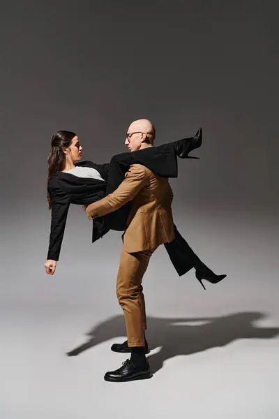 Homem de óculos e terno levantando mulher morena em saltos altos e traje formal em pano de fundo cinza — Fotografia de Stock