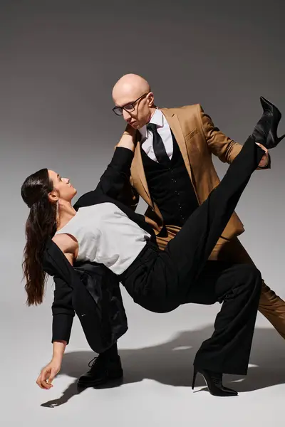 Homem de óculos e terno levantando mulher morena em saltos altos e traje formal em pano de fundo cinza — Fotografia de Stock