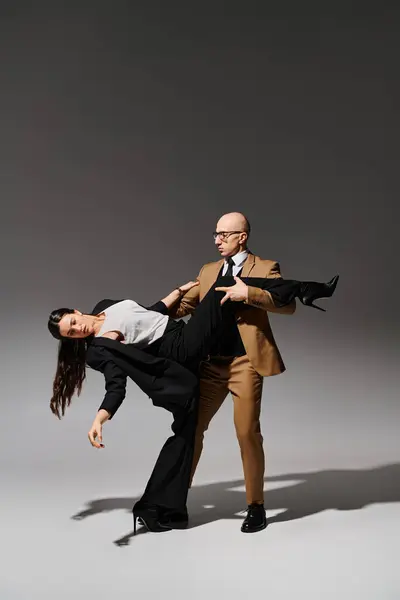 Man in glasses and suit supporting leg of brunette woman in high heels during dance on grey backdrop — Stock Photo