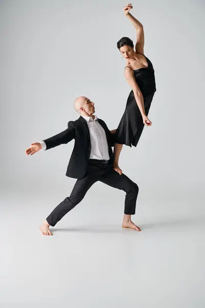 Barefoot dancer in black dress balancing gracefully on leg of male partner in grey studio — Stock Photo