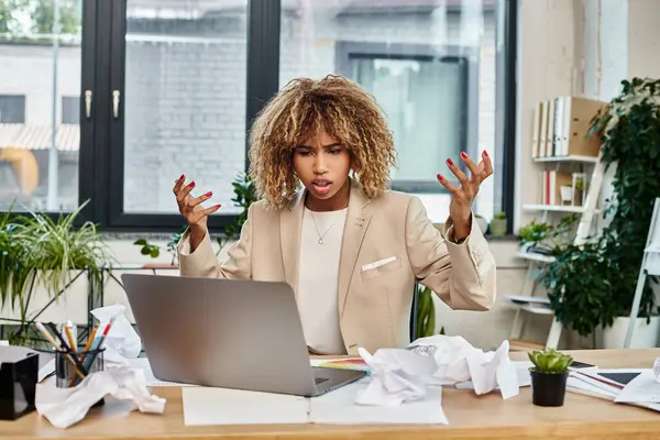 Irritado rizado africano americano mujer de negocios en su escritorio con ordenador portátil y papeles arrugados, estrés - foto de stock