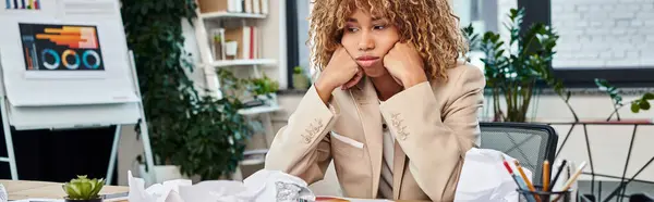 Cansada mujer de negocios afroamericana rizada en su escritorio con papeles arrugados, pancarta horizontal - foto de stock