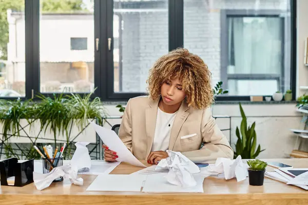 Encaracolado Africano americano empresária em sua mesa e olhando para documento perto de papéis amassados — Fotografia de Stock