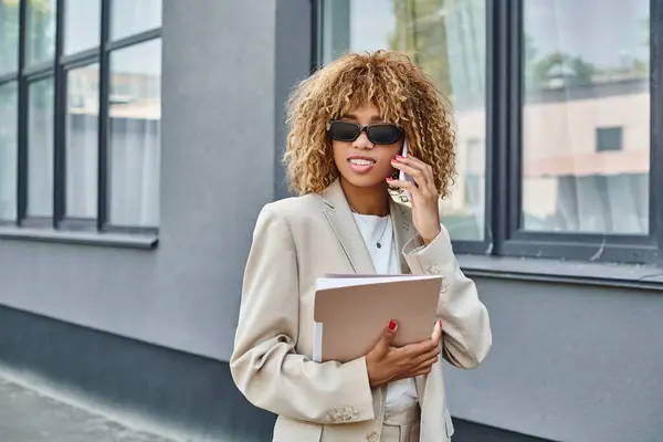 Alegre afroamericana mujer en traje y gafas de sol haciendo una llamada fuera de un edificio de oficinas - foto de stock