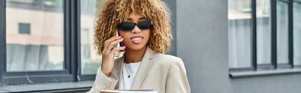 Cheerful african american woman in suit and sunglasses making a call near office building, banner — Stock Photo