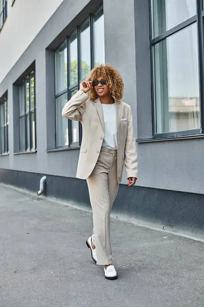Happy african american woman with braces in suit and sunglasses walking near office building — Stock Photo