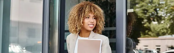 Happy african american businesswoman with curly hair holding folder and exiting building, banner — Stock Photo