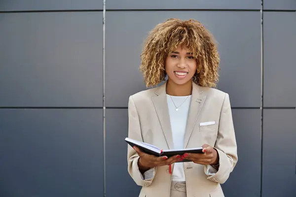 Mujer de negocios afroamericana positiva en ropa formal sosteniendo su cuaderno cerca del edificio de oficinas - foto de stock