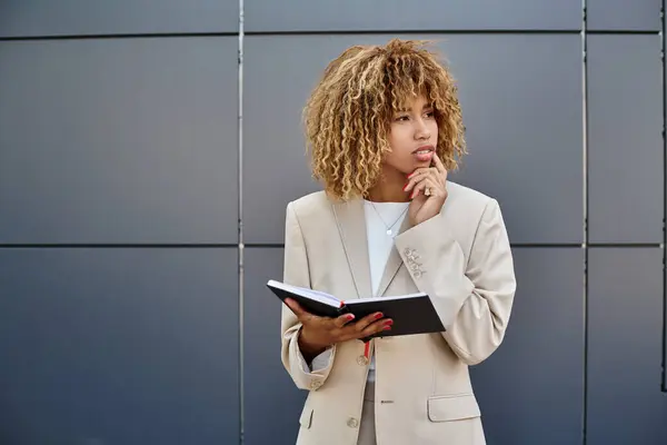 Pensativa mujer de negocios afroamericana en ropa formal sosteniendo su cuaderno cerca del edificio de oficinas - foto de stock