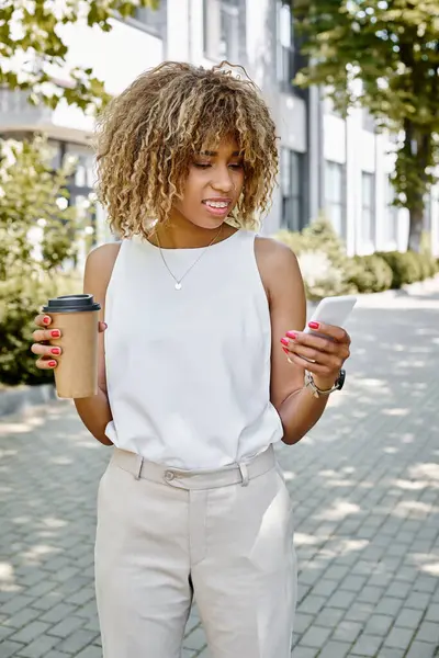 Allegra donna d'affari afroamericana in bretelle utilizzando smartphone e tenendo tazza di carta con caffè — Foto stock