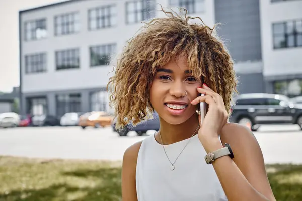 Retrato de mujer negra rizada radiante feliz que tiene llamada telefónica en el entorno urbano, teléfono inteligente - foto de stock