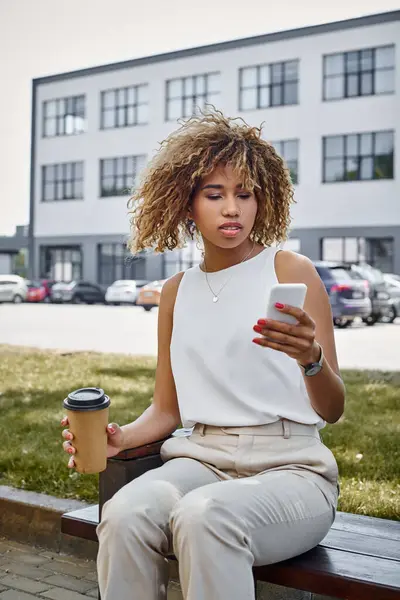 Femme d'affaires afro-américaine bouclée dans des bretelles à l'aide d'un smartphone et tenant une tasse de café pendant la pause — Photo de stock