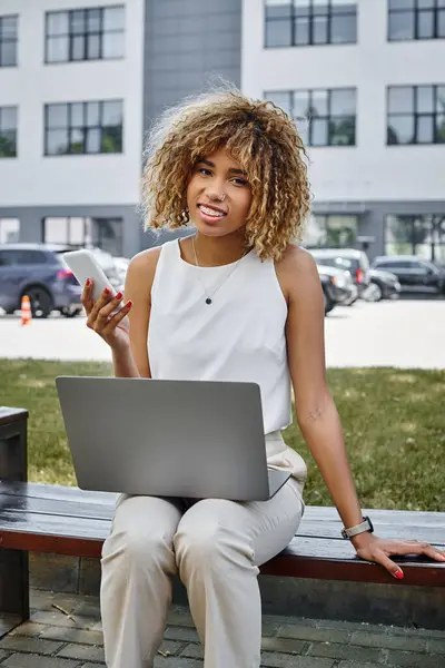 Happy african american freelancer with smartphone working on her laptop on bench in a park setting — Stock Photo