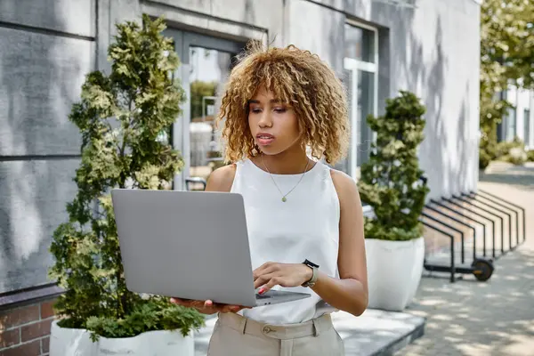 Ricci donna africana americana con bretelle in piedi con il suo computer portatile vicino edificio in calda giornata estiva — Foto stock