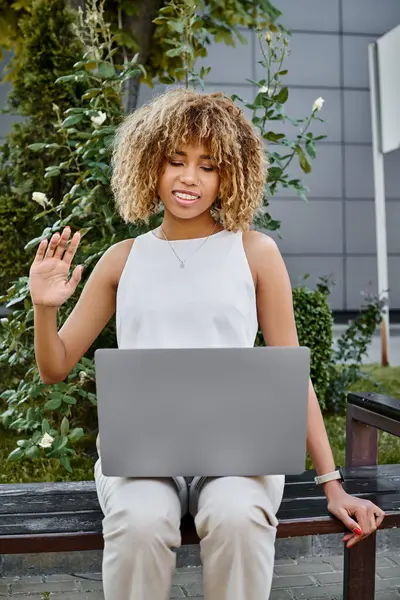 Happy curly african american freelancer having video call on her laptop and sitting on bench — Stock Photo